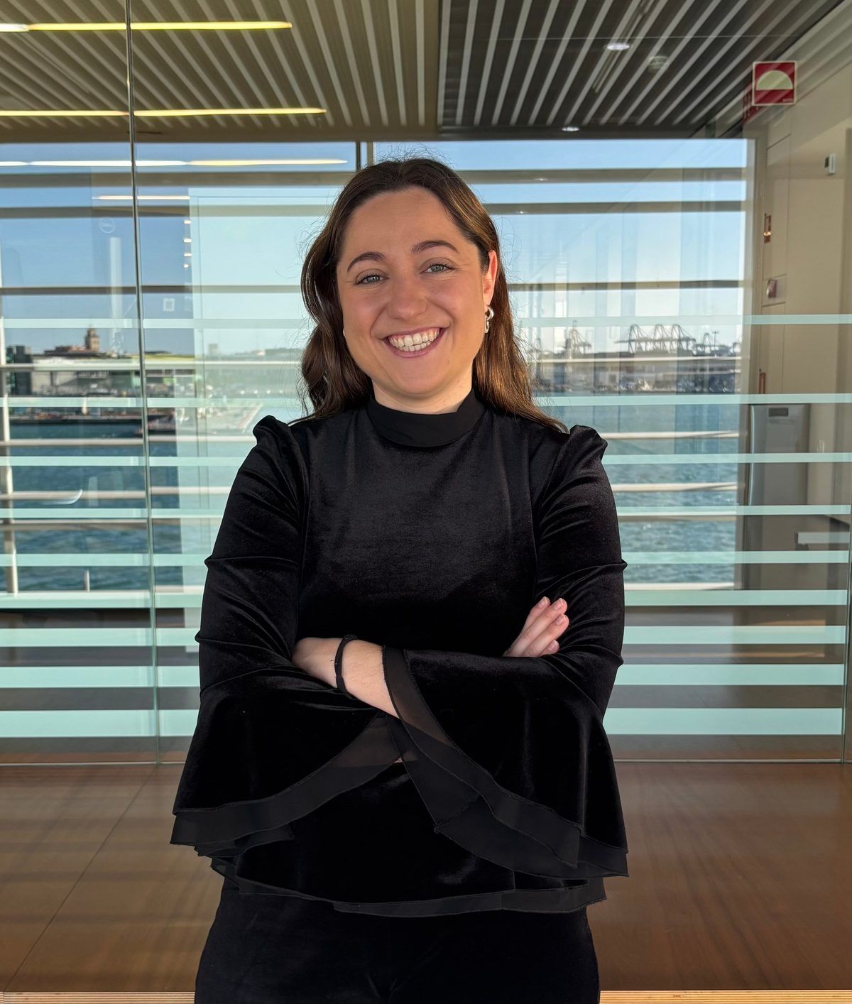 Retrato de Eneka Iribarren Parra, sonriendo con los brazos cruzados, con el puerto de fondo